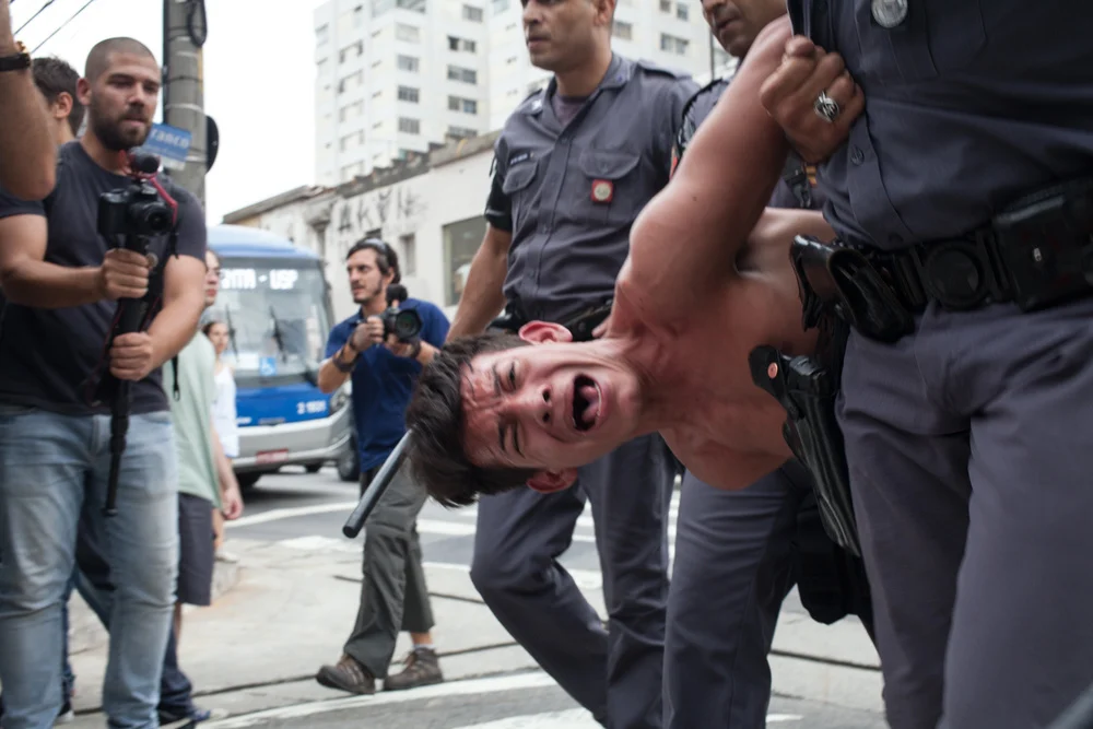 Foto de manifestação em São Paulo em 2015, mostrando um aluno sendo imobilizado por policiais.
